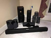 Photo shows a variety of black Sony speakers including a tall subwoofer, two bookshelf speakers, four satellite speakers, and a center channel speaker arranged on floor against wall.