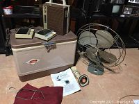 Vintage Electrophonic radio, Coleman cooler, rotary phone handset, vintage metal fan, and clothespin bag with instruction paper on floor