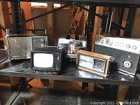Photo showing five vintage electronics: three radios, a toaster, and a small portable TV on a black shelf.