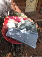 Photo showing red metal wheelbarrow with three gray gutter feet inside, viewed outdoors against brick wall and door.