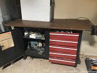 Full view of work table showing metal construction, worn wood surface, red drawers, open cabinet with contents, and side clamp