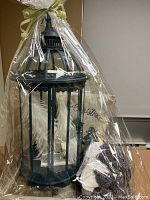 Gift-wrapped arrangement with dark teal metal lantern containing white feather Christmas trees, a cream pillow sham with script, and gray knitted throw blanket on a wooden tray