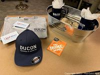 Galvanized metal tub containing two navy Ducon logo mugs with tissue, navy Ducon baseball cap, folded grey Ducon T-shirt, Home Depot and Boathouse gift cards displayed