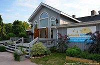 Exterior view of a gray lakeside building with ramp entrance and sign reading 'Restaurant on the Sandbanks - Waterfront Dining and Café', surrounded by planters and greenery