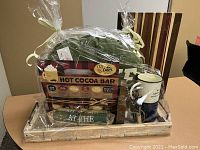 Gift basket wrapped in clear plastic featuring all included items: pillow, mugs, hot cocoa kit, wooden canoe decoration, tea towel, serving block, charcuterie board, all on a natural wood tray.