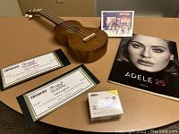 Photo of wooden ukulele, Adele 25 music book featuring Adele's face on cover, Korg ukulele tuner in box, and gift certificates for Novel Ideas and Frameworks