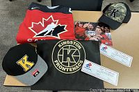 Lot arranged on table showing signed Team Canada jersey, signed photo, two ball caps, gold ticket vouchers in branded envelope and gift certificates.