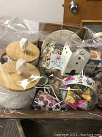 Photo of tea basket containing 2 white ceramic canisters with wooden lids, white ceramic teapot, colorful mugs, wooden house-shaped tea canister, decorative plate, and assorted tea bags. Items are wrapped in plastic.