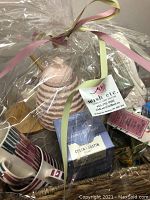 Photo of tea-time gift basket wrapped in clear cellophane with pink and green ribbons. Shows pink ceramic honey pot, Colin Justin tea box, striped mugs, and other assorted tea items tightly packed inside a woven basket.