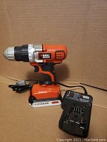 Black & Decker cordless drill standing upright with charger placed beside it. Orange and black color scheme, lithium 20V battery pack attached.