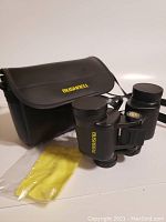 Bushnell binoculars with carrying case and yellow cleaning cloth laid out for display.