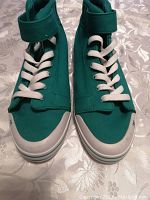 Front view of turquoise DC skateboard shoes with white laces and rubber toe caps on floral fabric background