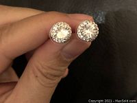 Close-up photo of two round stud earrings held between fingers, showing central clear stones with smaller stones arranged around the perimeter.