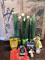 Overview of lot showing three artificial grass plants, Crayola clock, oriental-style wall hangings as background, lady figurine, and box with Chinese stress balls
