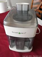 Front view of Vitamin Bar 2 juicer showing logo, feeding chute, and juice collector with measurement marks