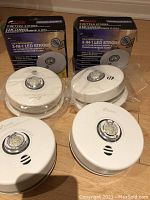 Four white Kidde 3-in-1 LED Strobe smoke and carbon monoxide alarms placed on floor with two still in sealed plastic bags and two out, with their original boxes behind them