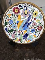 Front view of colorful ceramic plate featuring a bird and floral design with gold-trimmed scalloped edge on black display stand.