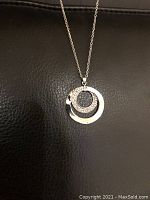 Photo of the necklace featuring a silver-tone circular pendant with smaller inner circle studded with crystals, hanging from a fine chain on black background.