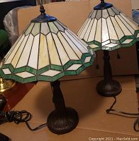 Pair of Tiffany style lamps with stained glass shades in green and cream tones, metal bases, and cords shown on cardboard background.