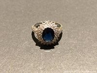 Front view of the ring showing the oval blue center stone surrounded by clear stones in a halo arrangement with additional clear stones on the band.
