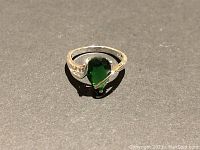 Sterling silver ring with a large green teardrop-shaped gemstone and small clear accents on the band shown against a dark background under bright light.