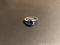 Front view of the silver-tone ring featuring an oval blue stone in the center, surrounded by smaller clear and blue stones, lying on a dark surface.