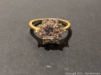 Top view of gold tone ring with central large clear gemstone and surrounding smaller stones, showing details and sparkle.