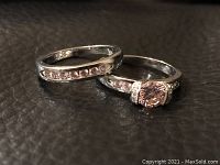 Two silver rings shown on black surface, one with row of small clear stones, other with large pink center stone