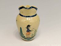 Front view of ceramic Quimper pitcher showing Breton man with fishing rod motif and greenery, cream colored glaze with blue trim.