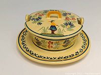 Front view of the butter crock with lid and affixed underplate showing the Breton man motif and floral decoration.