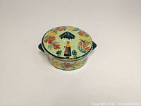 Top-front angle view of the covered ceramic box showing the Breton woman under umbrella and floral pattern on lid and body.