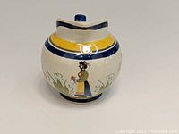 Photo front view of the HenRiot Quimper creamer showing Breton woman holding flowers and grass motif on white background with yellow and dark blue bands near the rim and base.