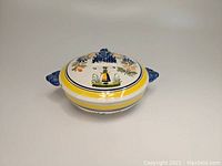 Front angle of HenRiot Quimper covered footed serving bowl displaying lid with Breton woman and floral motifs, side handles, and footed base.