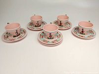 Full shot of the 5 pink pottery teacups and 6 saucers arranged in a row showing hand-painted Breton figures and floral motifs.