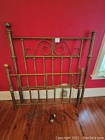 Full view of the antique brass bed frame including headboard and footboard with decorative swirls and vertical bars, set against a red wall and wooden floor.