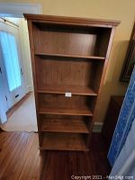 Photo showing full view of five-shelf wooden bookcase with natural medium brown wood finish, positioned indoors near a door and a mattress leaning on the right side.