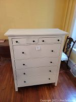 Photo shows a white painted Ikea chest of drawers with six drawers; two smaller ones on top and four larger drawers beneath. The drawer knobs are round and black. The chest stands on the floor in a room with wood flooring.