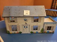 Front view of the closed sheet metal dollhouse showing painted siding, windows, door, roof, and floral landscaping at the base.