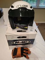 Front view of HJC XL motorcycle helmet on top of its original box with pair of black and orange utility gloves in front.