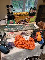 Full lot of outdoor camping gear including tent, stove, lanterns, ropes, and accessories arranged on a table and in boxes.