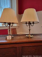 Pair of brass candle-style lamps with cream pleated lampshades on a wooden surface near a window.