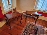 Photo of a pair of matching vintage mahogany tables with pull-out shelves extended. Tables have cabriole legs and rectangular tops, displayed in a room with hardwood floors and a window with lace curtains.