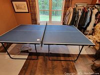 Full view of the assembled blue ping pong table with net, black metal legs, and wheels indoors
