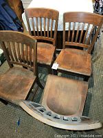 Four mid-century wooden office chairs arranged closely together on a carpeted floor. Chairs have vertical slatted backs and contoured wooden seats with visible wear.
