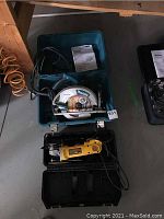 Wide view showing the Makita circular saw in teal case above and DeWalt angle grinder in black case below, cords visible, instruction manuals included