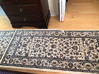 Top view of one rug placed on wood floor near dark wood furniture, showing floral and vine detailed design in beige, dark blue, and light brown colors.