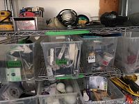Wide view of metal shelving with clear plastic bins containing assorted hardware, plumbing parts, and supplies along with light bulbs and tape.