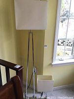 Tall floor lamp with brushed metal triangular base and rectangular white shade placed in corner near window and stairs.