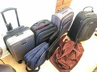 View of four pieces of luggage including gray hard shell with USB port, black and navy soft-shell suitcases, brown leather shoulder bag, two black leather paper carriers, and a bag of electric adapters.