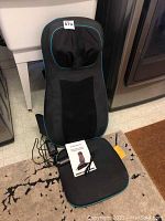 Black massage cushion with turquoise piping laid on floor, shows head and back padding, power cord, and user manual on seat section.